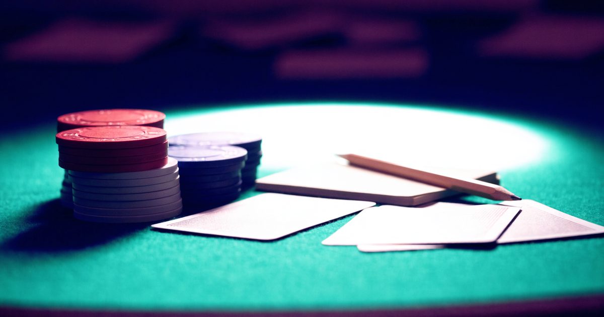Stacks of poker chips, face-down playing cards and a notepad with a pencil on a green felt poker table under a soft overhead light.