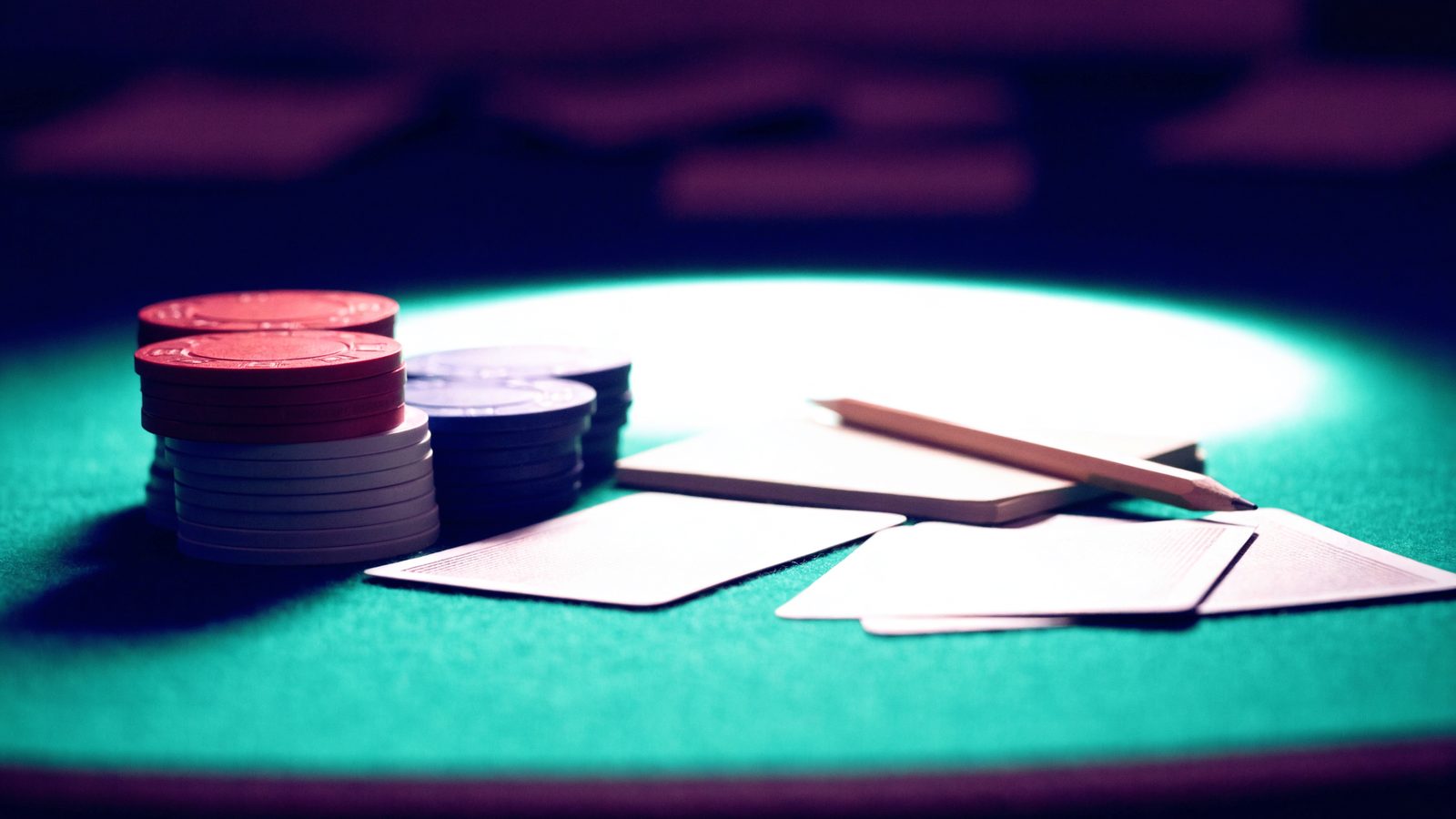 Stacks of poker chips, face-down playing cards and a notepad with a pencil on a green felt poker table under a soft overhead light.